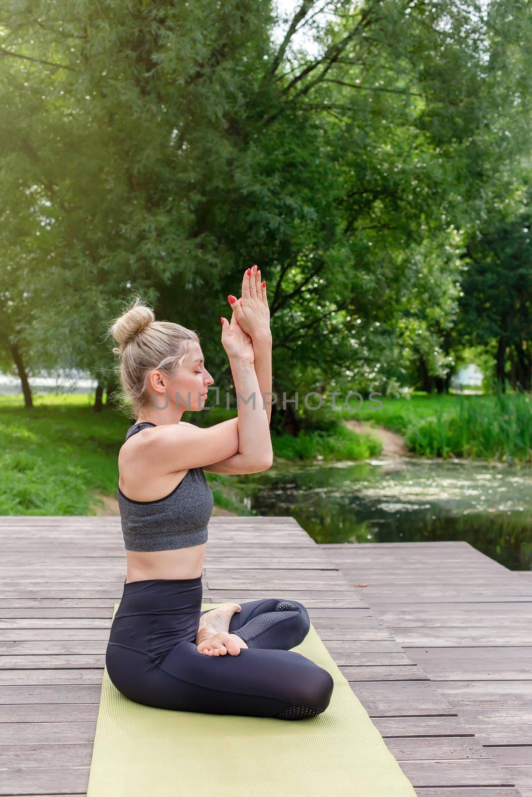 A beautiful woman in a gray top and leggings, sitting on a wooden platform by a pond in the park in summer, does yoga on green mat . Vertical. Copy space