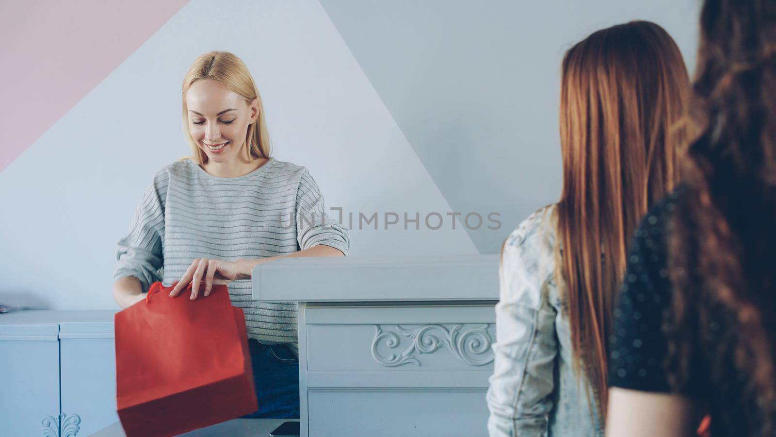 Friendly female cashier is taking garment from customer standing in line folding and packing clothes in paper bag to buyer by silverkblack