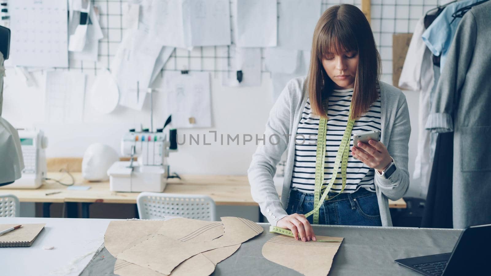 Clothing design entrepreneur is checking paper sewing drafts and looking at smartphone. She is examining every cutout carefully and thinking about future garment. by silverkblack