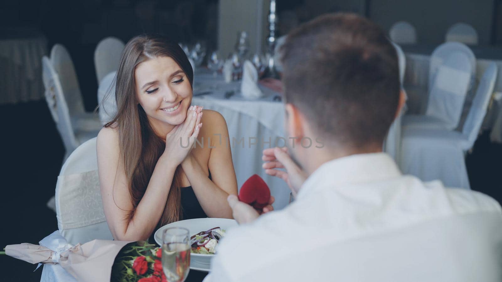 Handsome guy is proposing to surprised pretty woman, then putting engagement ring on her finger and kissing her hand during romantic date in restaurant. by silverkblack