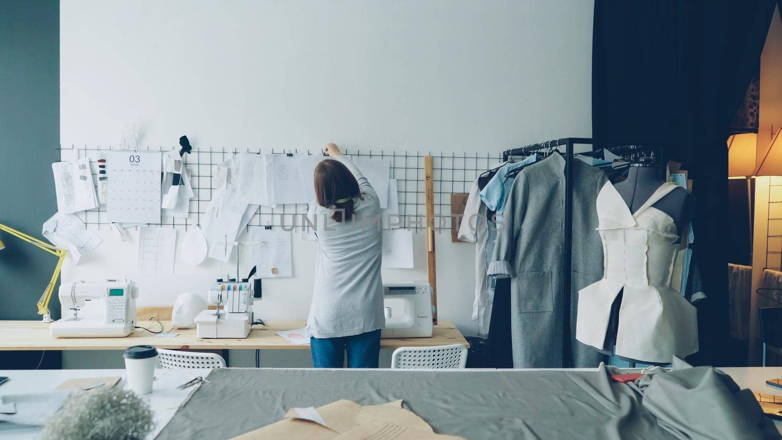 Female fashion designer is choosing and hanging clothing sketches to wall for her newest collection, then watching them carefully. Light fabrics, clothes hanging and sewing items are visible.