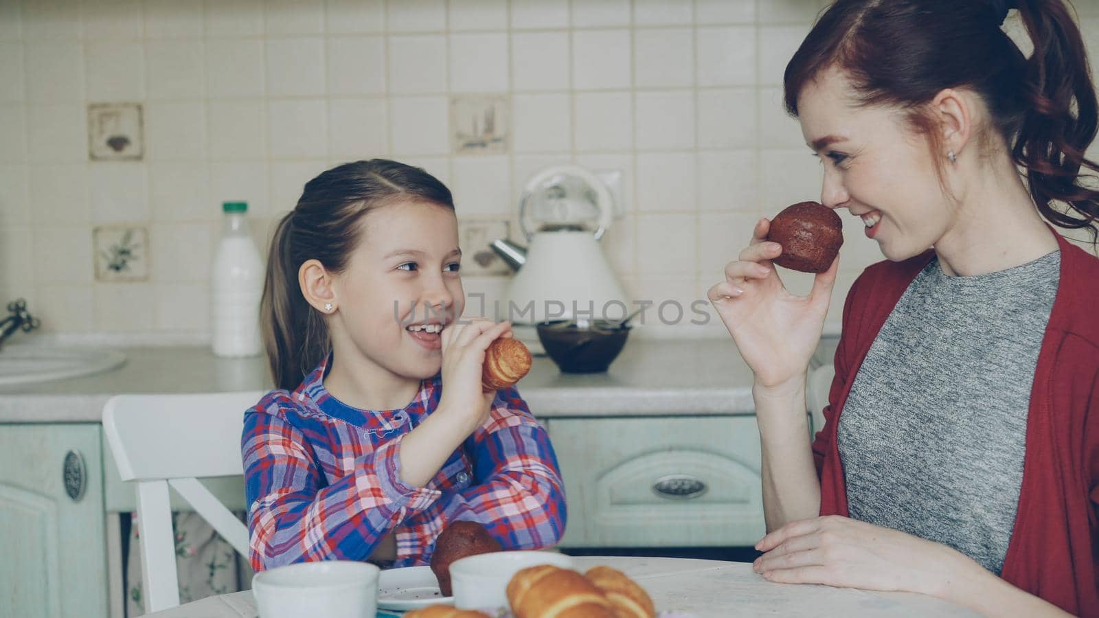 Happy young mother and cute daughter having breakfast eating muffins and talking at home in modern kitchen. Family, food, home and people concept