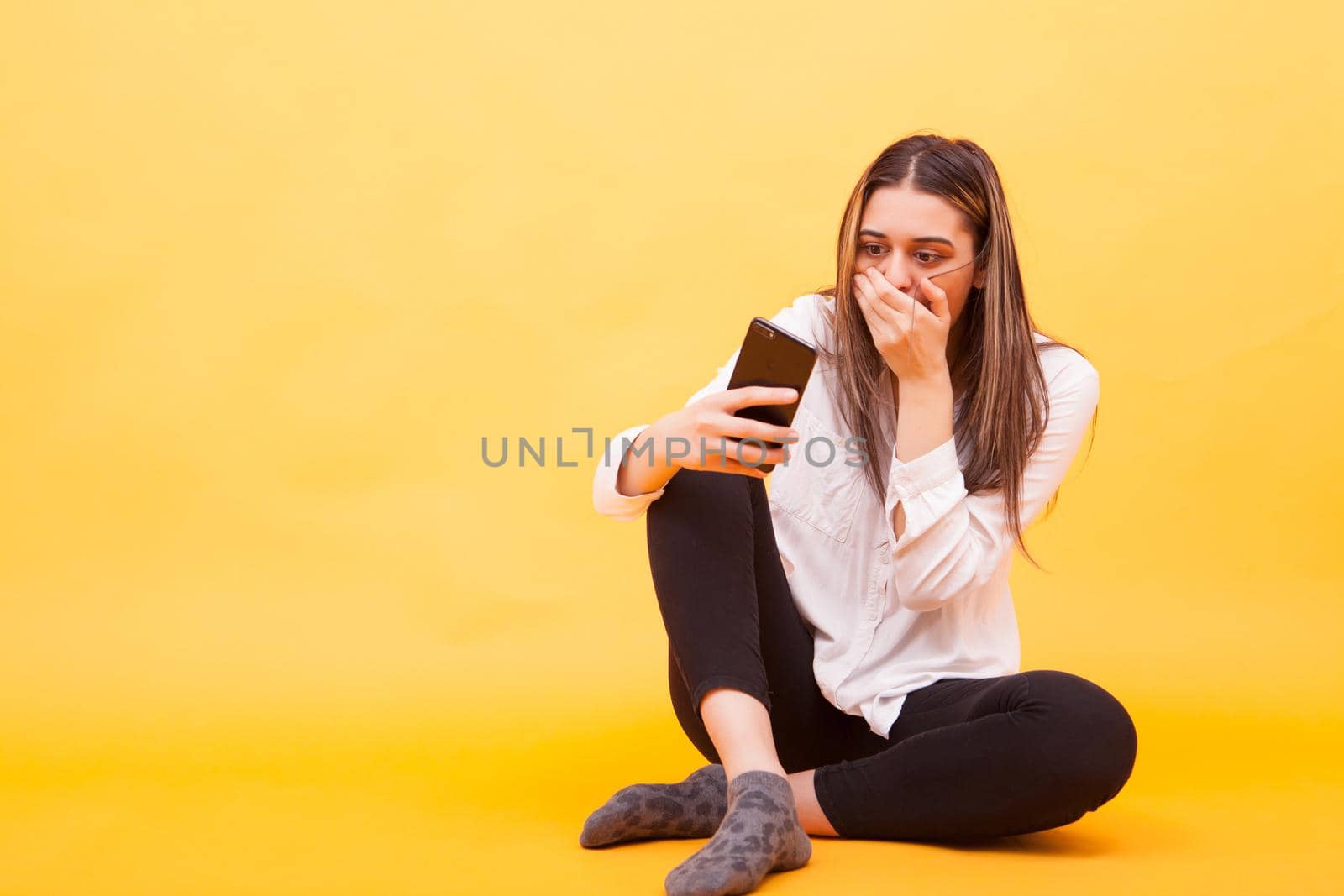 Girl looking shocked at her phone while sitting down over yellow background. Facial expression