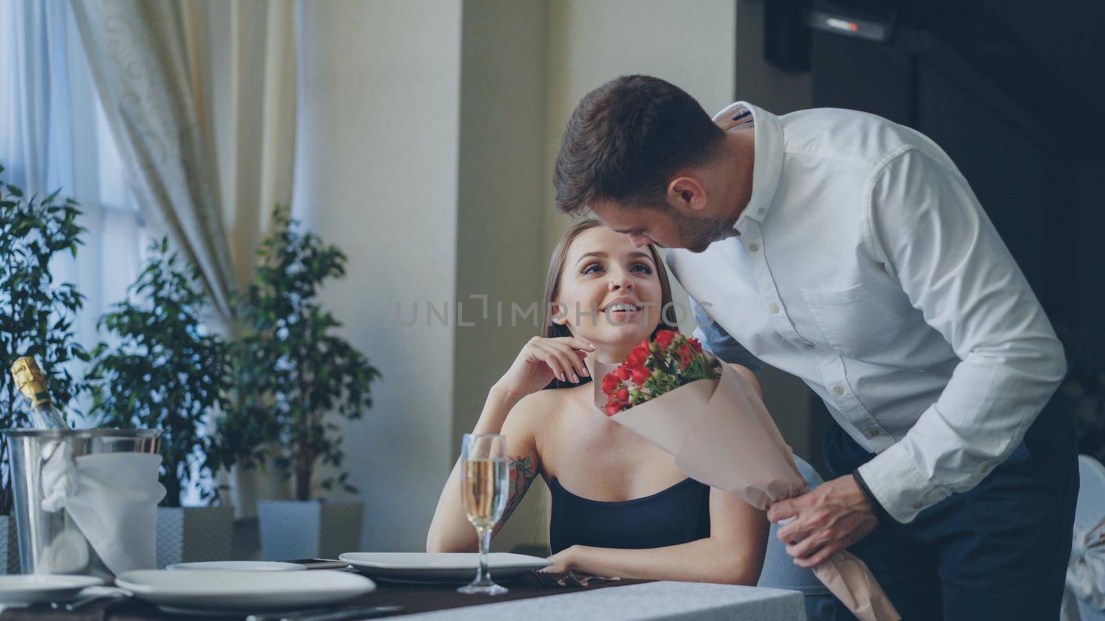 Beautiful woman is waiting for her boyfriend in restaurant, he is coming and giving her flowers, kissing her. Happy girl is smelling flowers and smiling with pleasure. by silverkblack