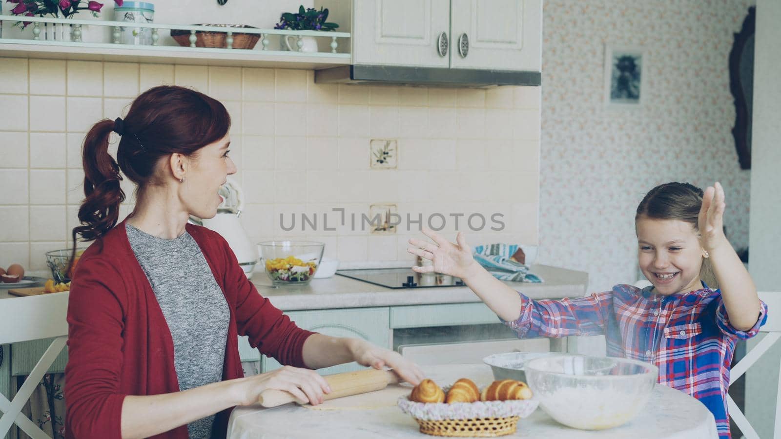Young mother and cute daughter cooking together talking in the kitchen on weekend. Little girl clapping hands with flour and laughing. Family, food, home and people concept by silverkblack