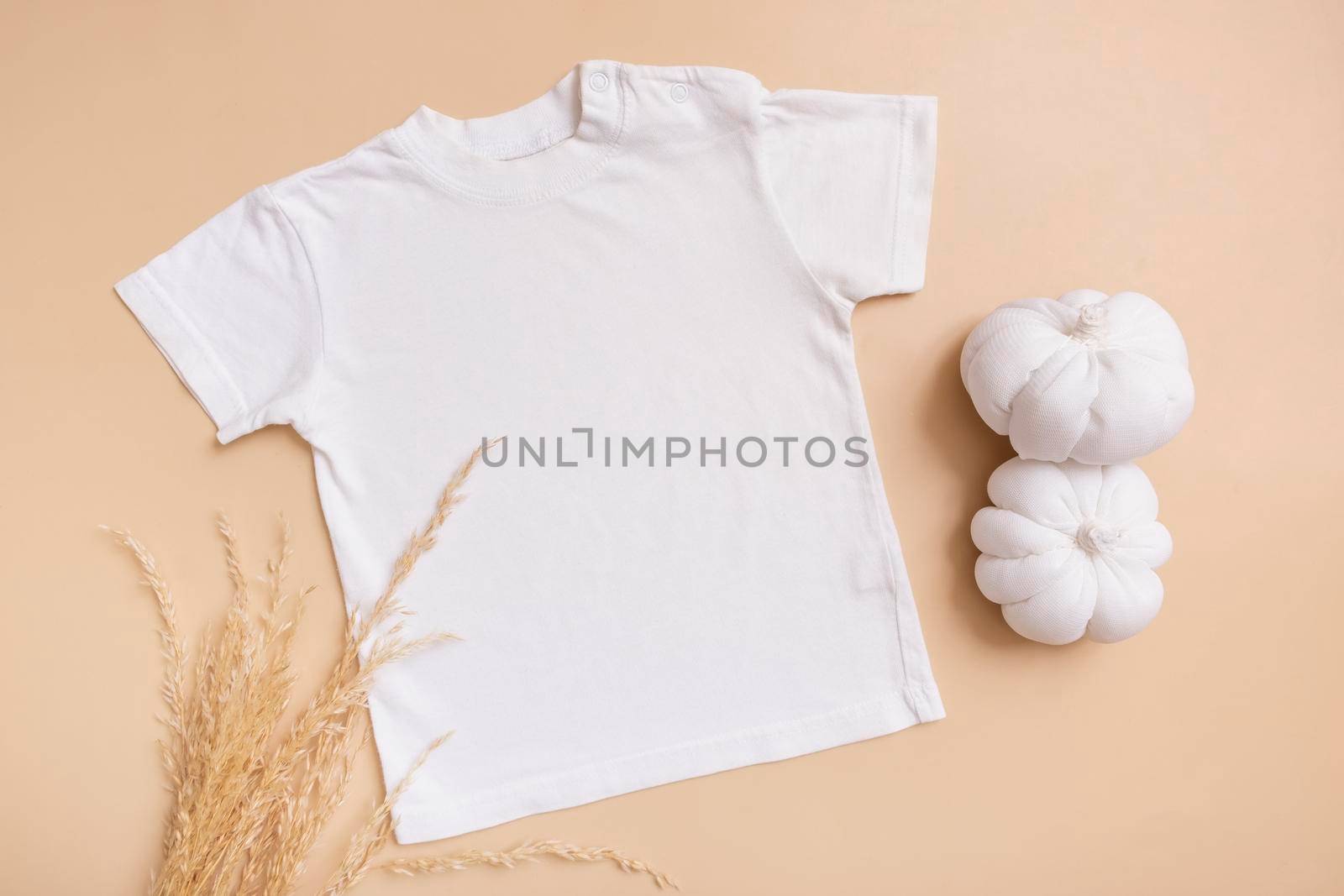 White baby t-shirt top view. Mock-up for logo, text or design on beige background. Flat lay child clothes with decorative pumpkins.