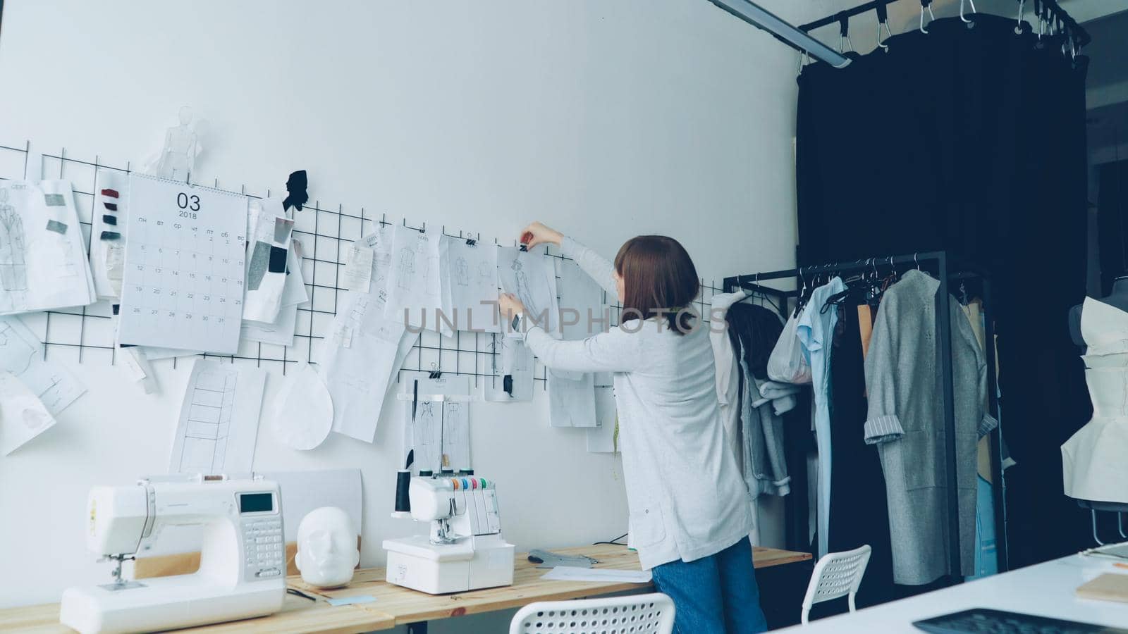 Female clothing designer is taking sketches from studio table and putting them on wall with other drawings of trendy women's garments. Creative thinking concept.