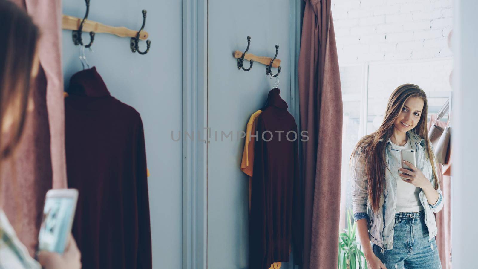 Pretty girl is making mirror selfie with smartphone while standing in fitting room in clothes store. She is posing, turning and touching her hair with lovely smile. by silverkblack