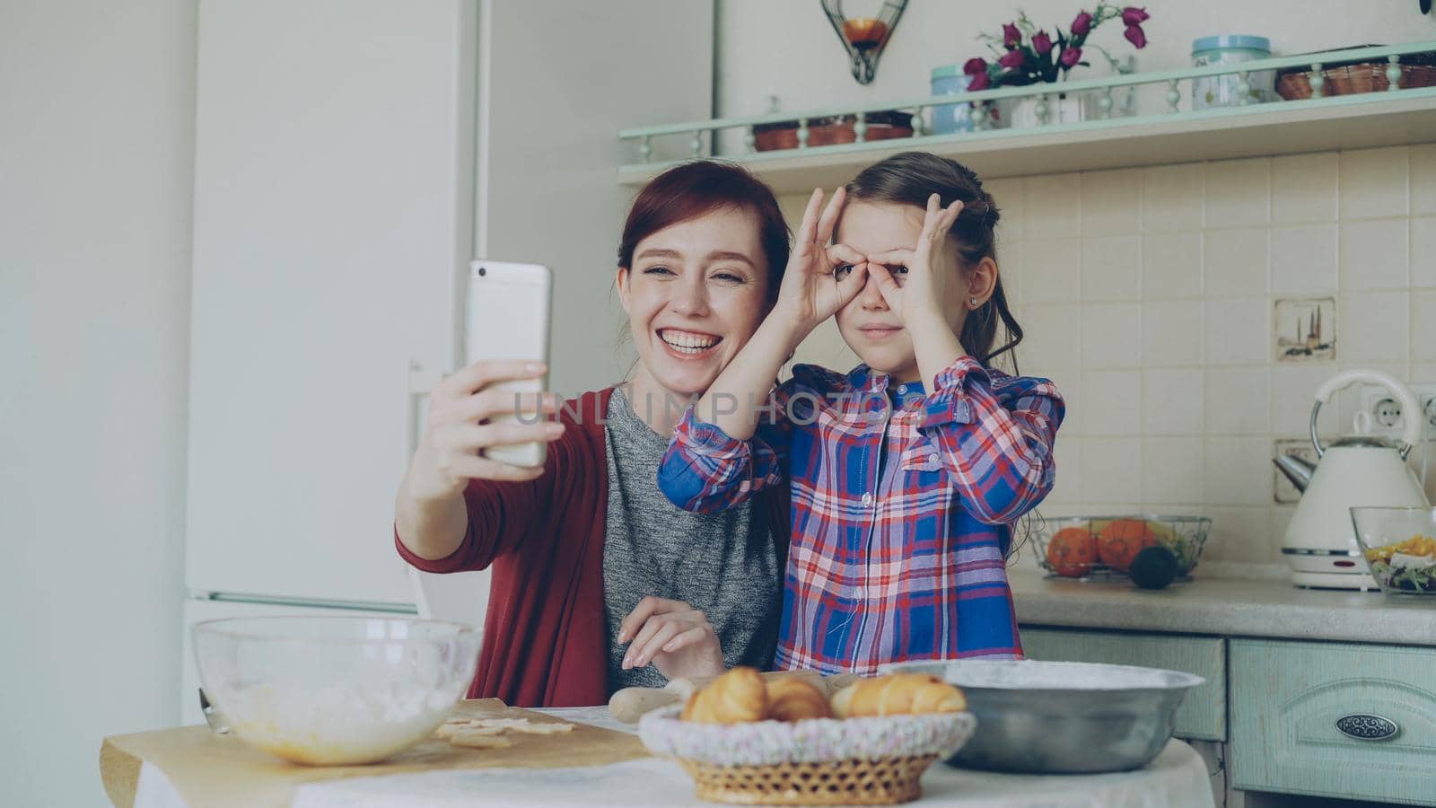 Smiling mother together with funny daughter taking selfie photo with smartphone camera making silly face while cooking at home in kitchen. Family, cook, and people concept by silverkblack