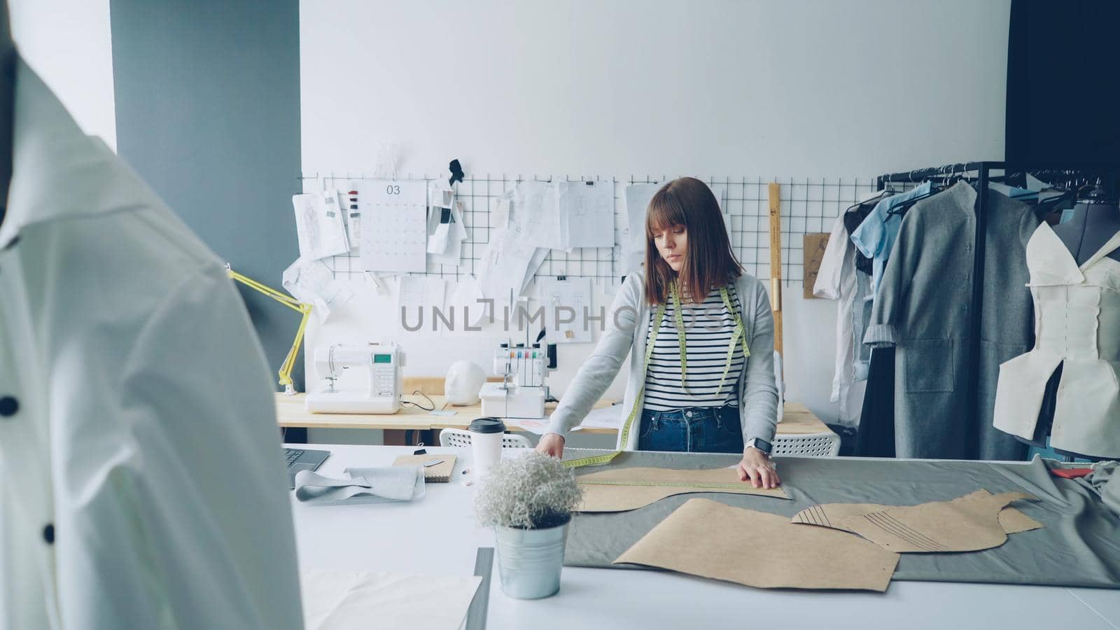 Young attractive female designer is checking her paper templates with measuring tape. Professional creative clothing designing concept. Studio is light and modern with many sewing items.