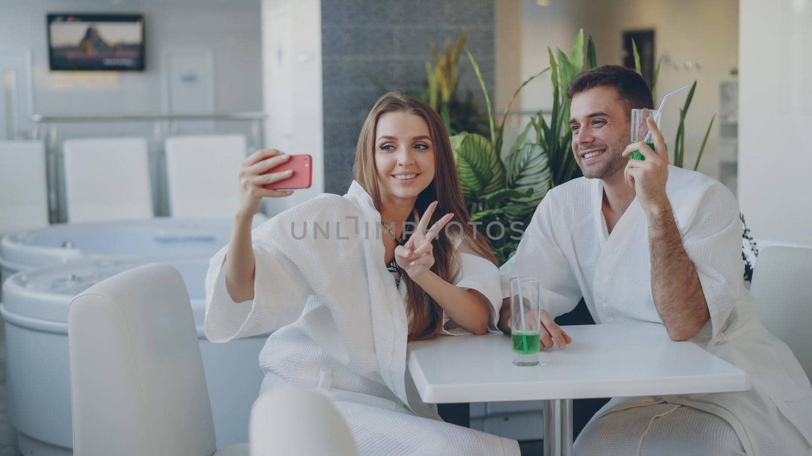 Pretty cheerful girl and her loving boyfriend are taking selfie with cocktail glasses using smartphone while relaxing in spa salon. They are smiling and posing looking at camera.