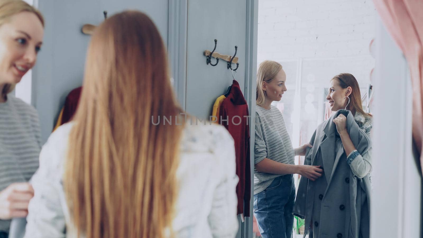 Pretty young woman is checking fashionable coat in fitting room with her friend helping her to appraise garment. They are talking, gesturing and looking at clothing. by silverkblack