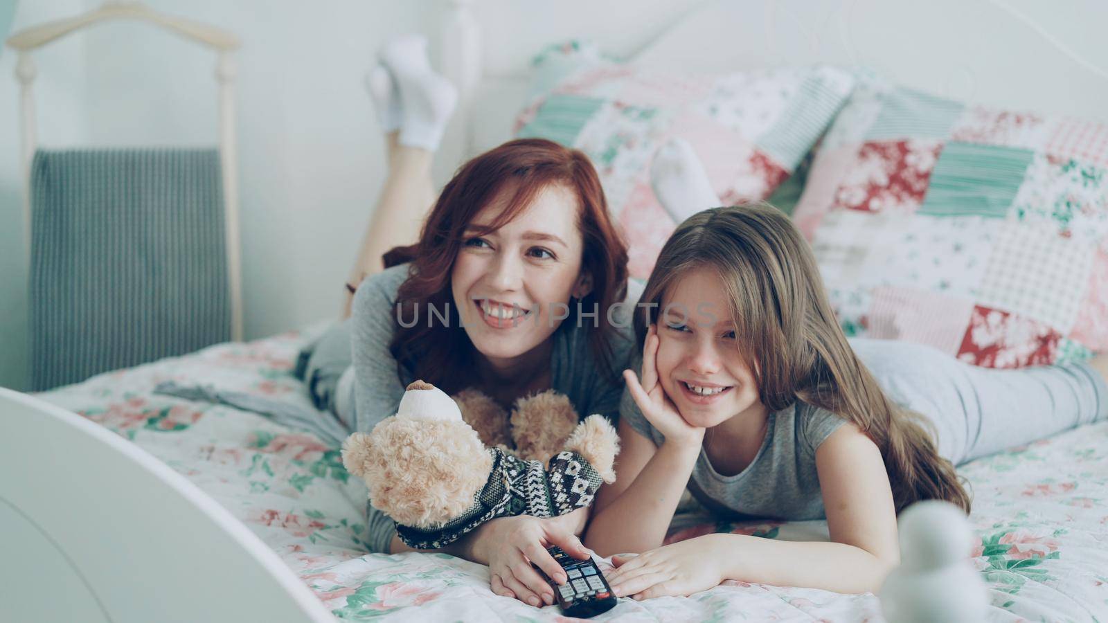 Laughing happy mother and cute daughter watching funny cartoon movie on TV while lying on bed at home in the morning and have fun by silverkblack