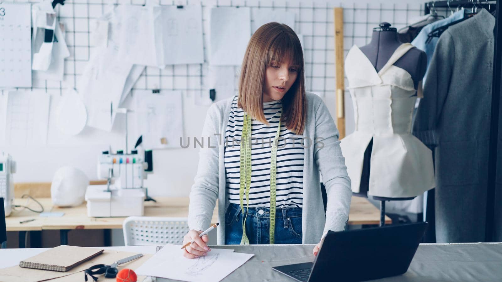 Beautiful fashion designer is drawing clothing sketches for her new clothes collection and working with laptop. Light studio with garments on rails and drawings on wall in background.