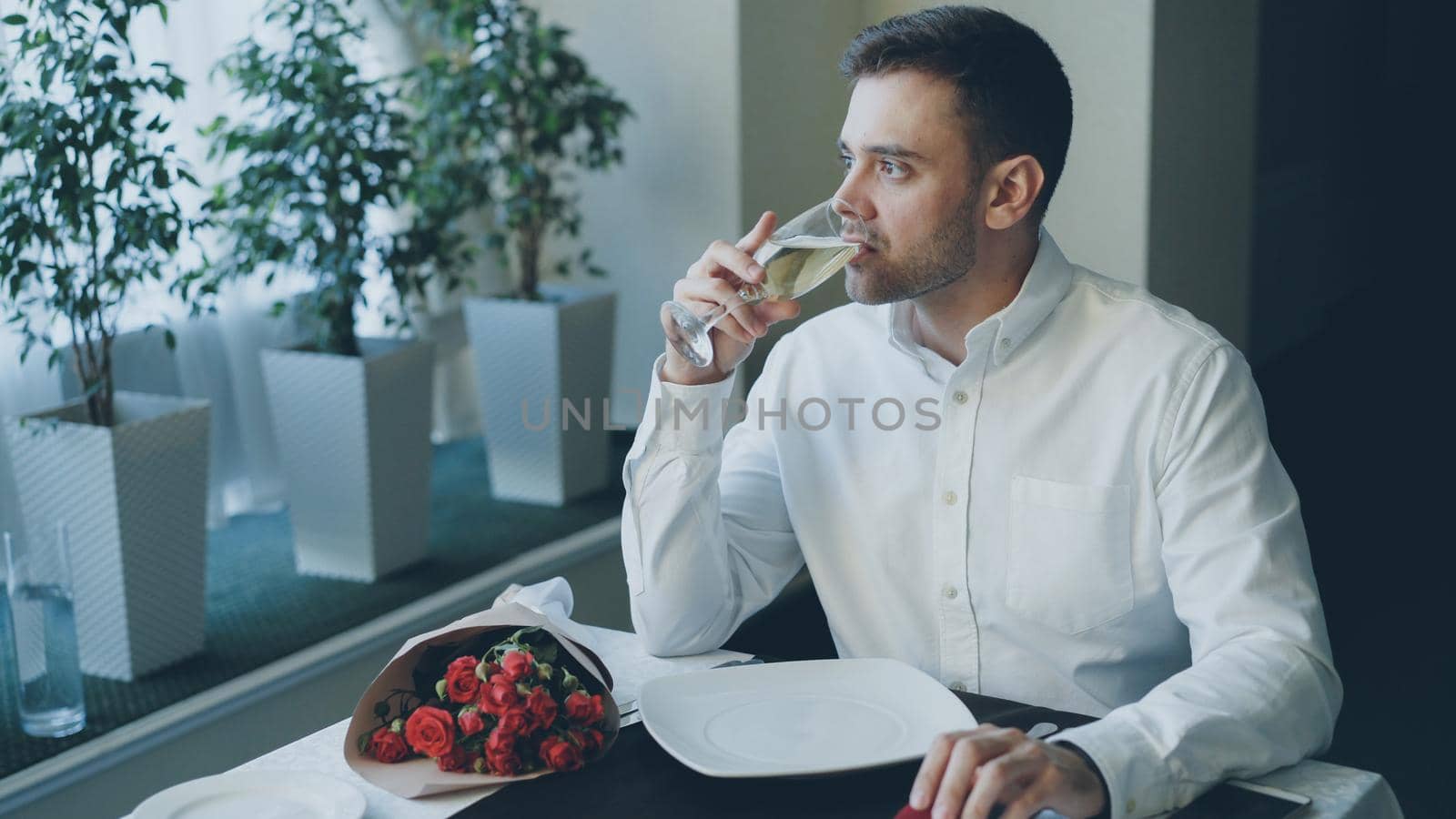 Nervous handsome man is sitting alone at table in restaurant, drinking champagne and waiting for his girlfriend, then leaving. Bunch of red roses, jewelry box and smartphone are visible.