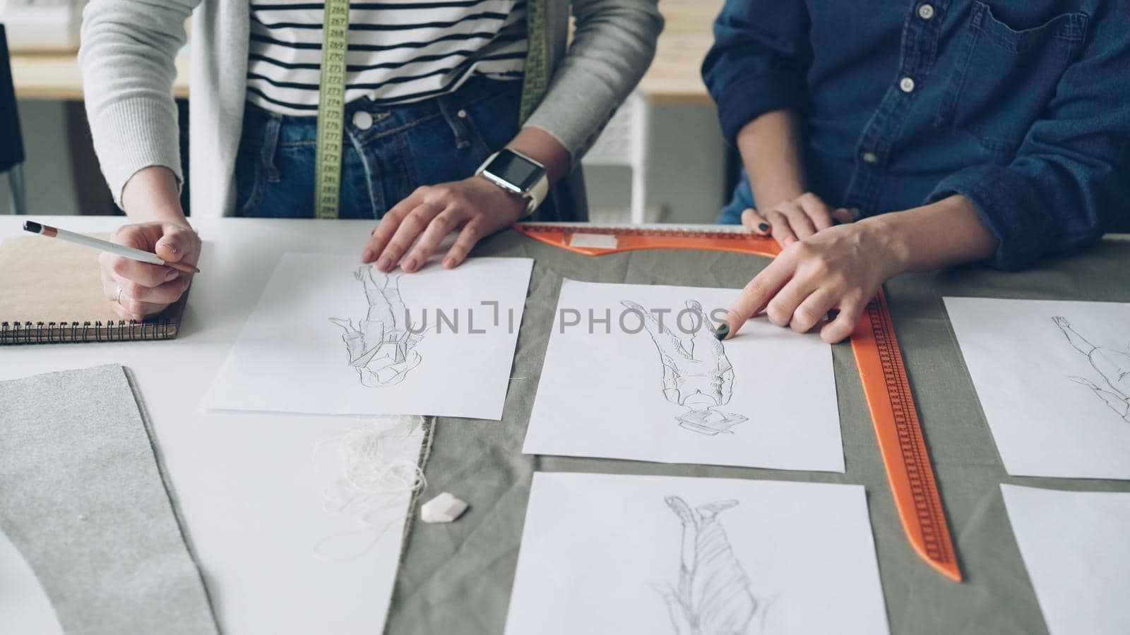 Close-up shot of female designers' hands choosing sketches for new clothing collection. Drawings of trendy garments are on table together with fabric and sewing accessories.