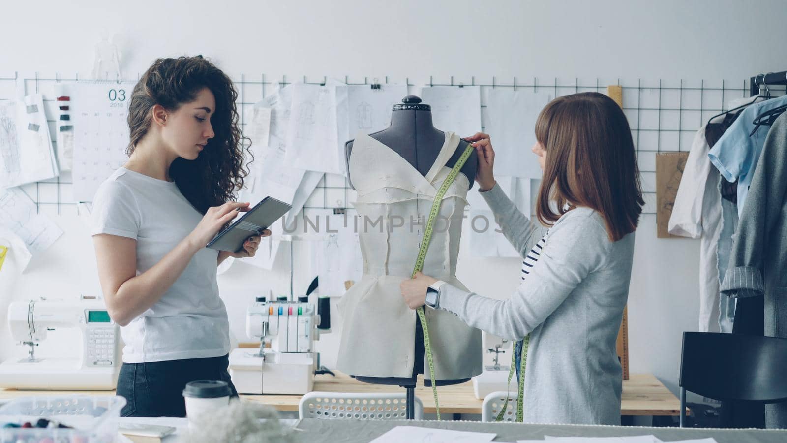 Professional seamstress is measuring patterns on mannequin with measure-tape while her coworker is working with tablet to write down measurement data. Light modern workshop in background.