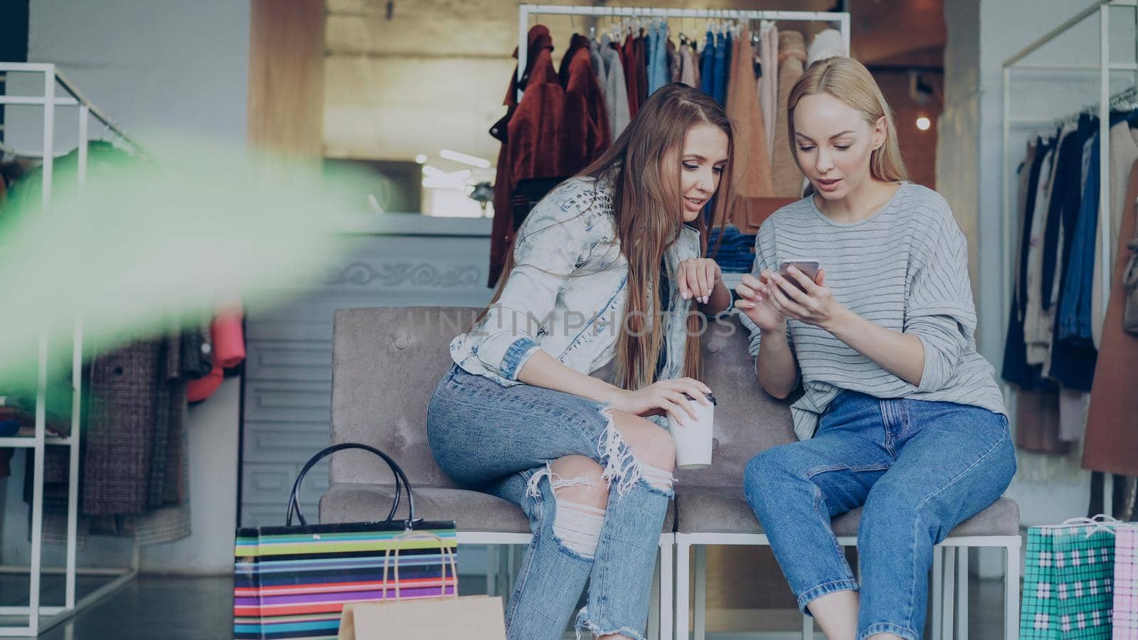 Attractive female friends talking and checking smartphone while sitting is modern shopping center. Colourful women's clothing, shelves, hangers and paperbags in background.