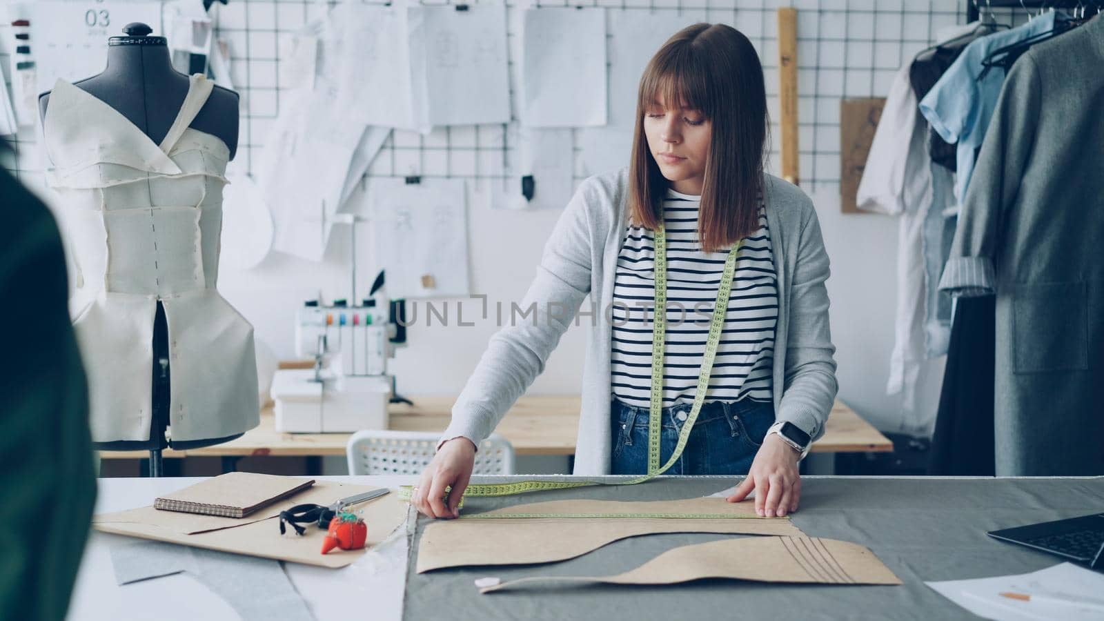 Young pretty seamstress is measuring clothing patterns with tape-measure at studio table while working in her modern tailor's shop. Getting ready for sewing clothes concept.