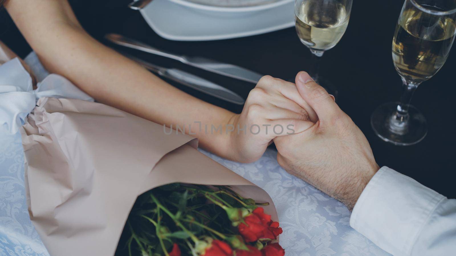 Close-up shot of male hand holding and squeezing female hand on table with champagne glasses and flowers. Romantic relationship, love and fine dining concept. by silverkblack