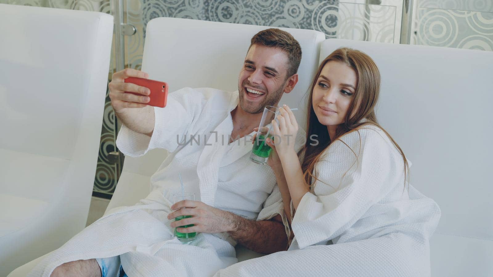 Attractive loving couple is taking selfie with cocktail glasses using smartphone while relaxing in spa salon. They are smiling, talking and posing looking at camera.