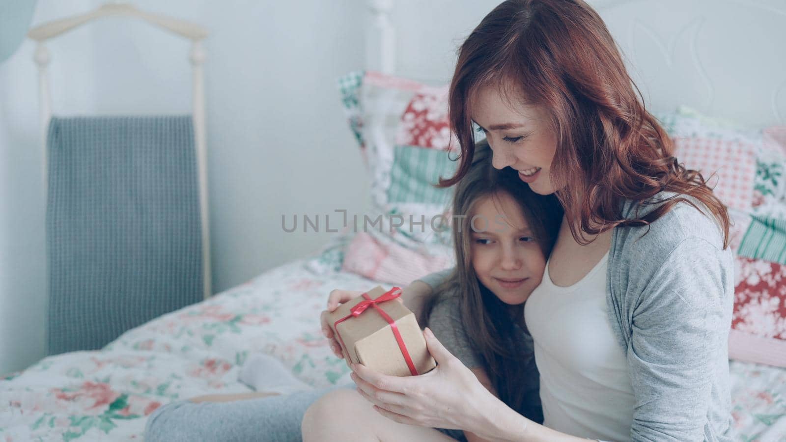 Happy younf mother kissing her cute daughter presenting gift box on celebration sitting on bed in light cozy bedroom at home