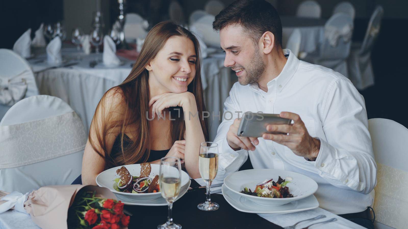 Happy attractive young people are watching smartphone together, smiling and talking while having dinner in restaurant. Modern technologies and romance concept. by silverkblack
