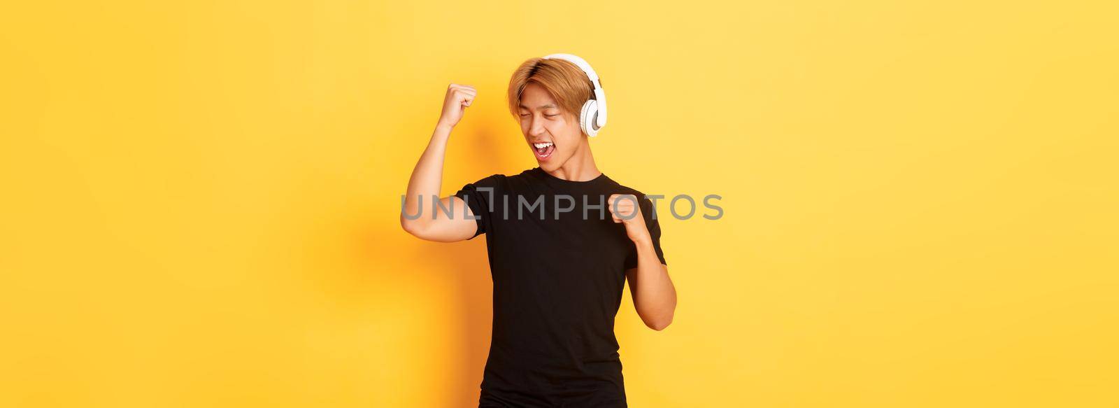 Carefree attractive asian guy with blond hair, singing along and dancing as listening to music in wireless headphones, standing yellow background.