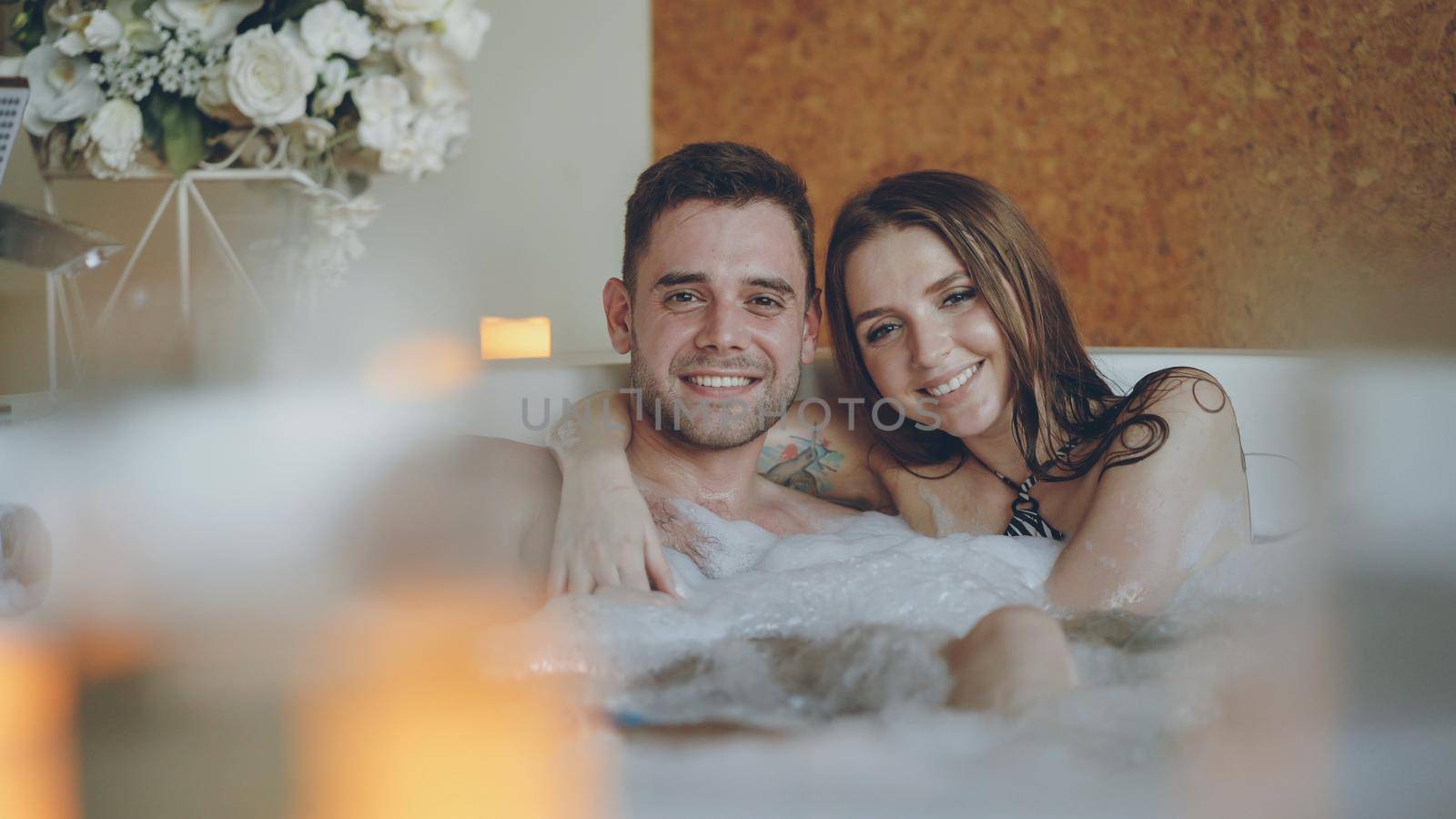 Portrait of good-looking young lovers hugging in jacuzzi, smiling and looking at camera. Burning candles, beautiful flowers and bubbling water with foam is visible.