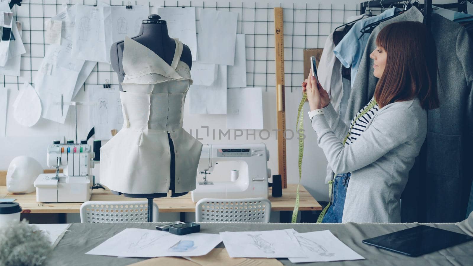 Self-employed female tailor taking photos of it with smartphone. Nice workshop with sewing machines, sketches and garment in background.