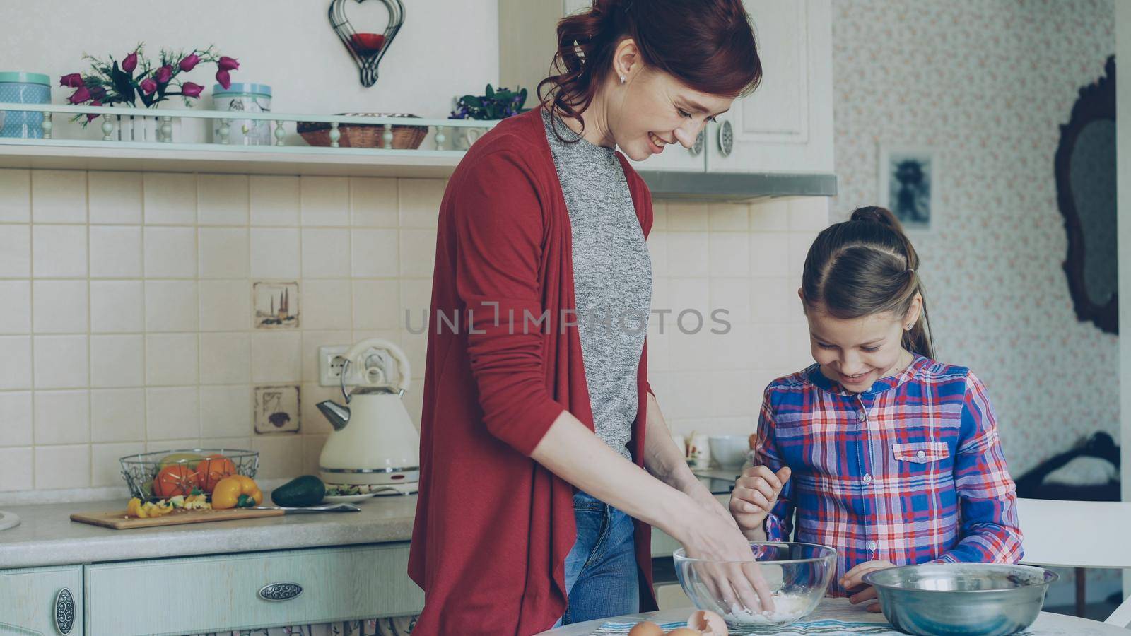 Little cheerful girl helping her mother in the kitchen mixing dough for cookies. Family, food, home and people concept