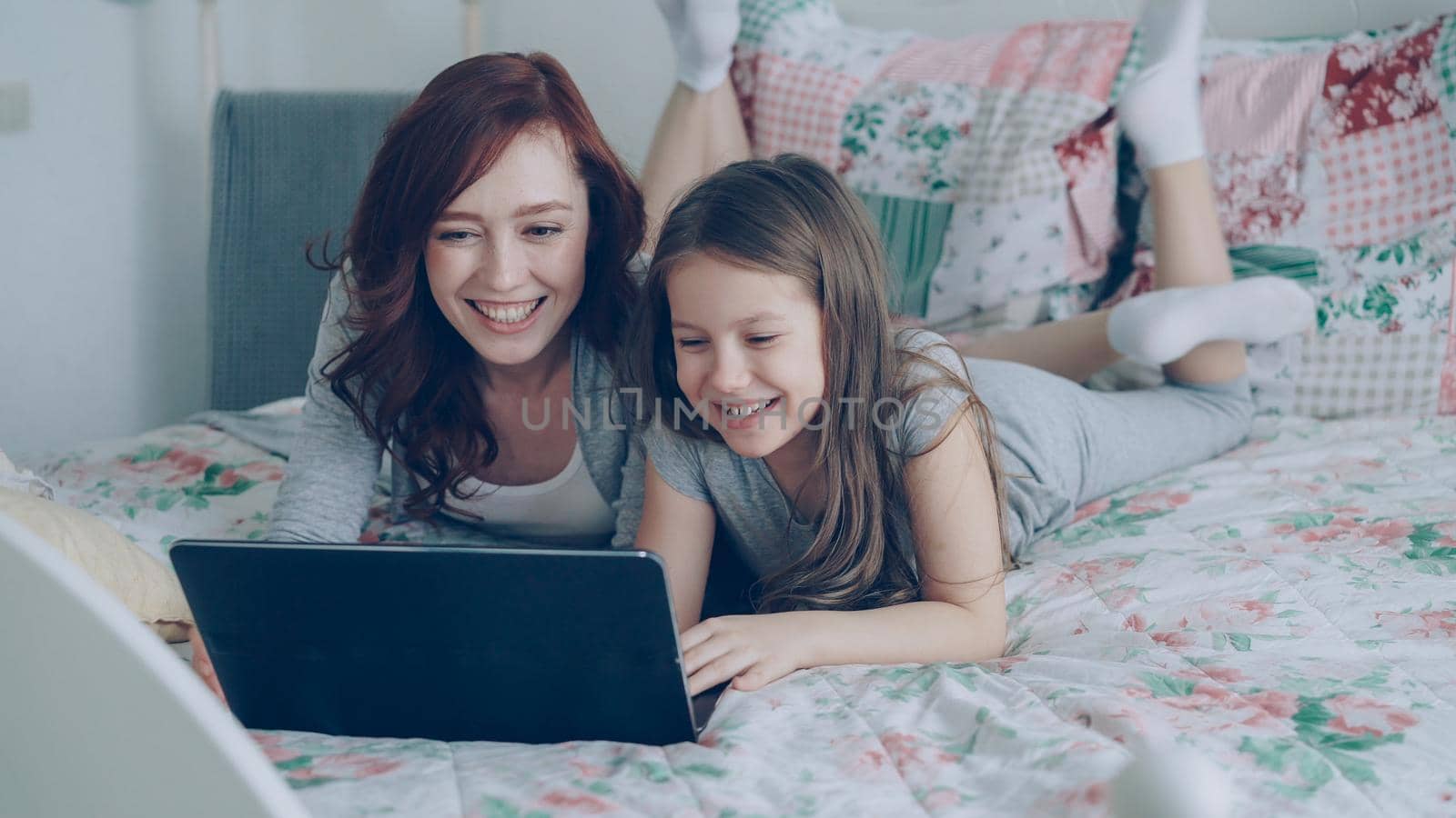 Happy cute daughter and young mother have online video call with father or grandmother using laptop computer lying on bed in cozy light bedroom at home.