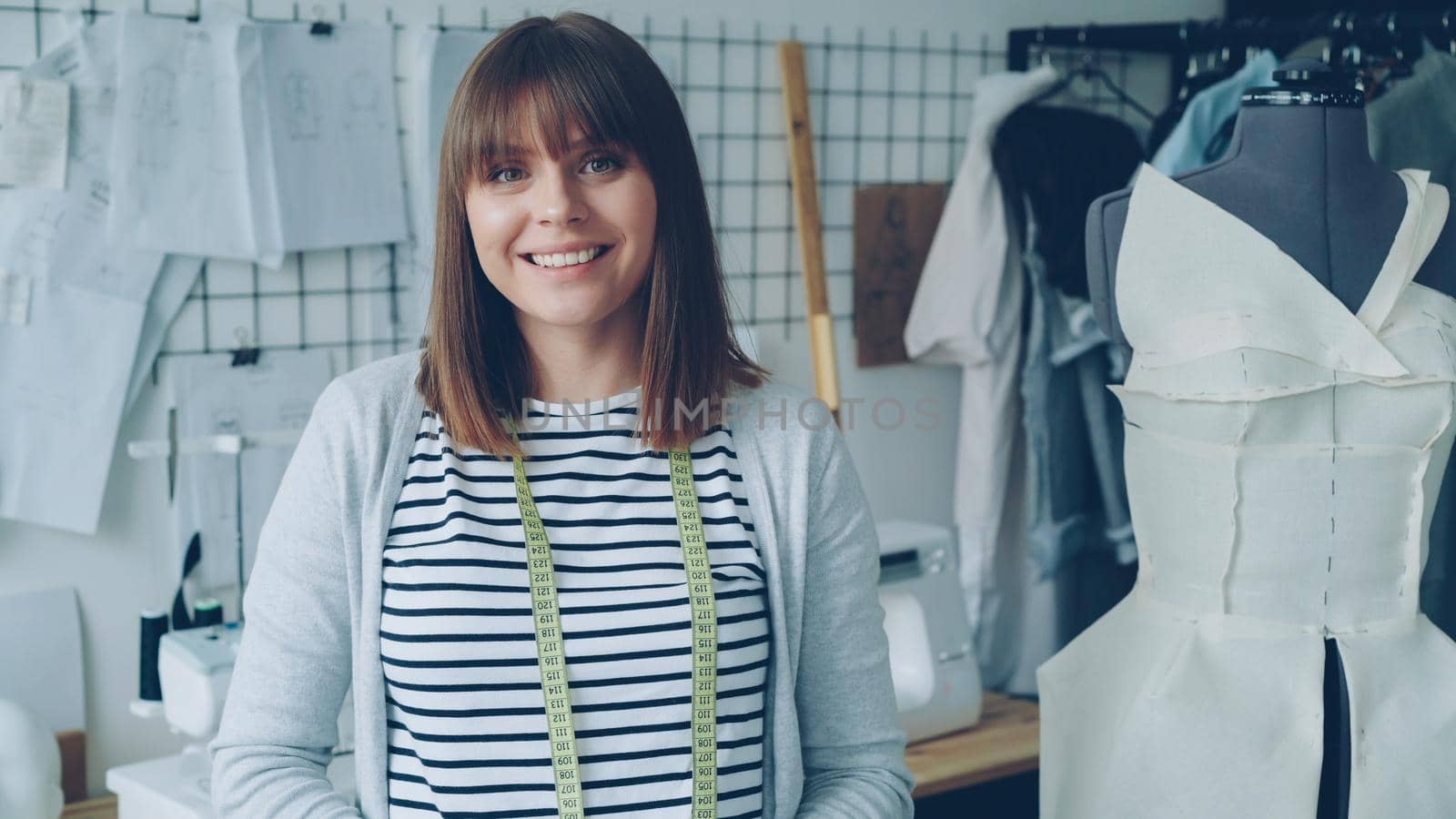 Portrait of attractive young woman clothing designer with brown hair looking at camera. Smiling woman is standing beside dummy, light modern tailoring workshop in background. by silverkblack