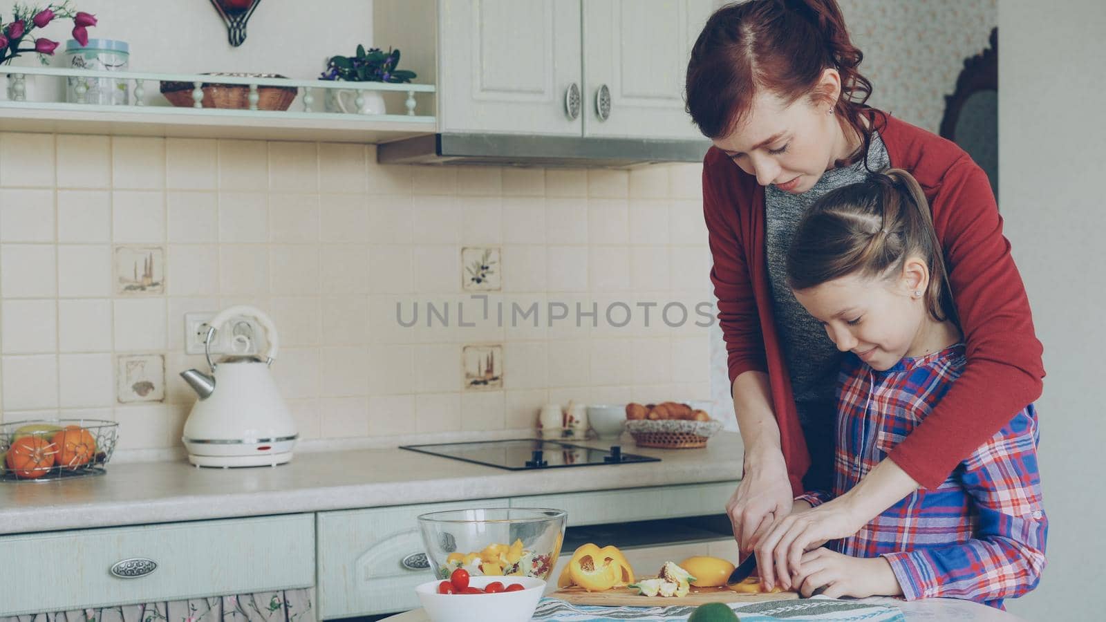Young pretty mother teaching her cute daughter to cut vegetables properly. Little girl cooking together with loving mom at home in modern kithcen by silverkblack