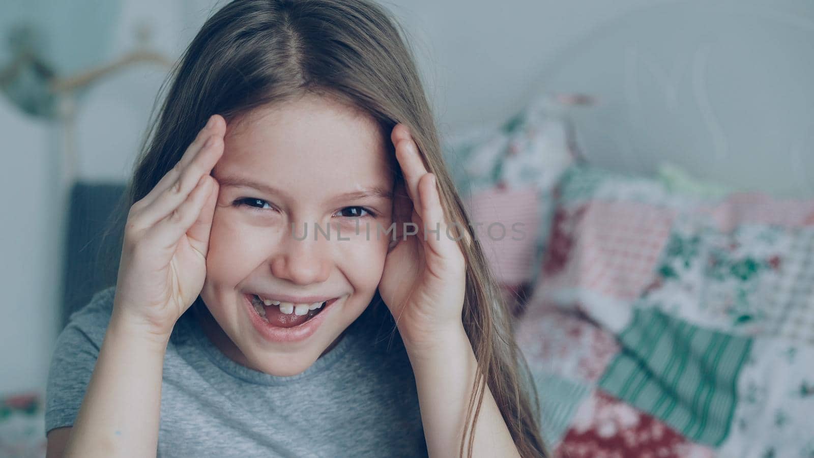 Close-up of Cute little funny girl covers her face and open hands smiling and grimacing looking into camera sitting in bedroom at home by silverkblack