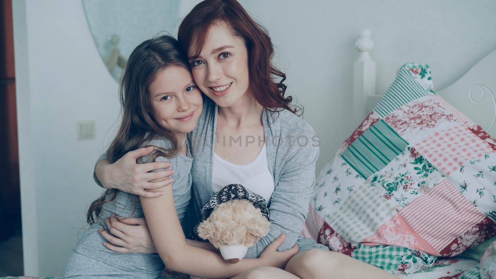 Portrait of adorable smiling girl embracing her happy mother and looking at camera together while sitting on bed in bright cozy bedroom at home