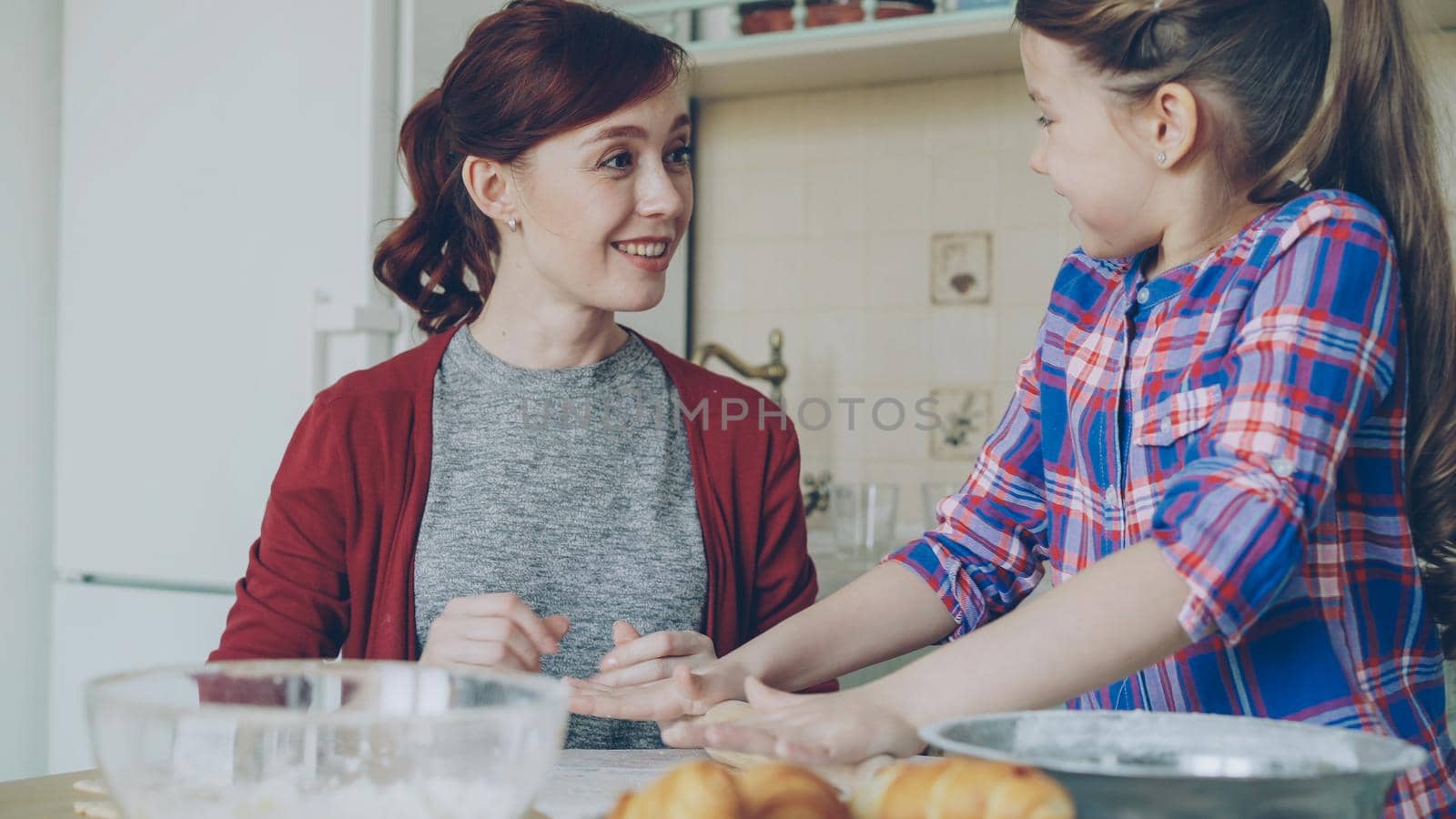 Cheerful mother and little cute daughter rolling dough while cooking in the kitchen on weekend. Family, food, home and people concept