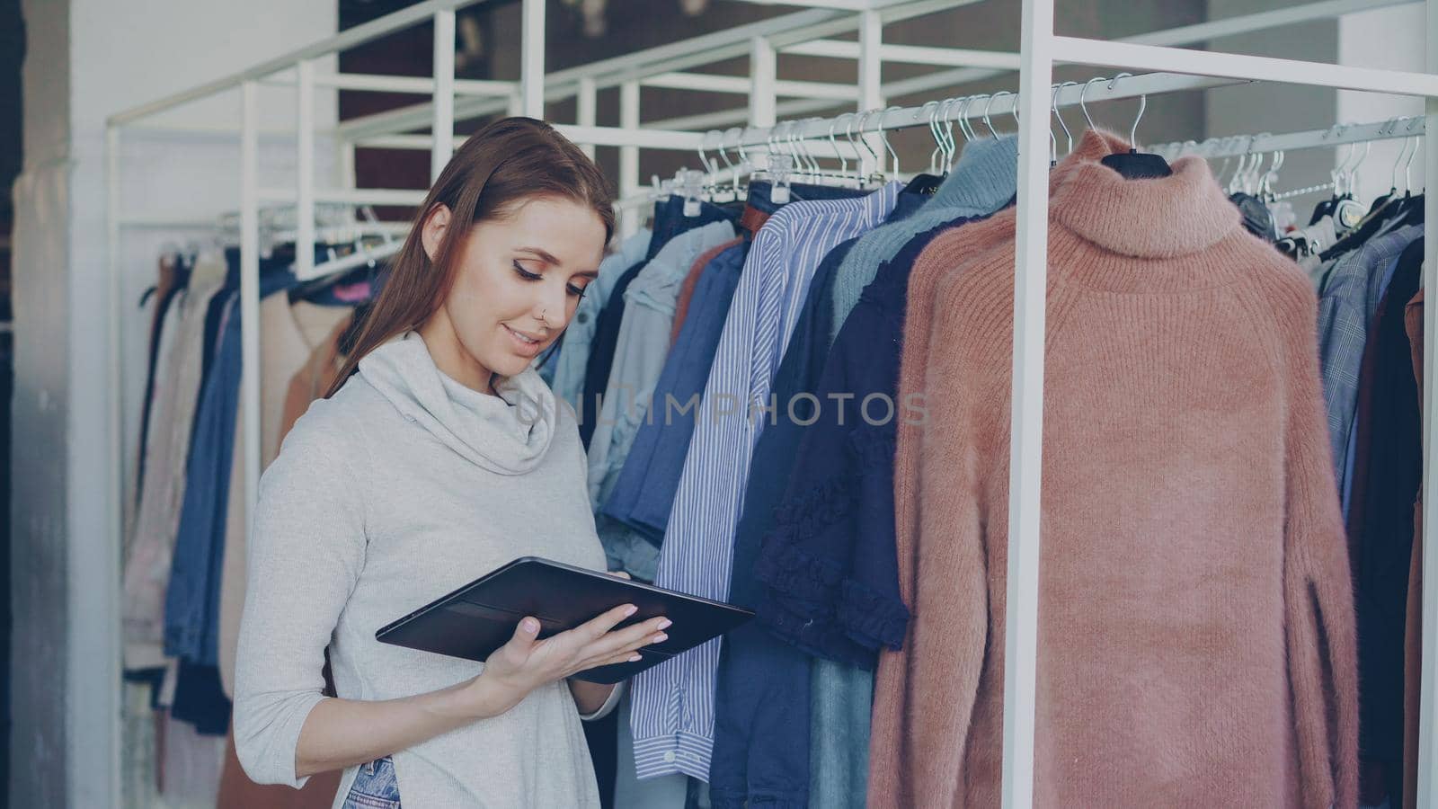 Owner of women's clothing shop is checking and counting garments on rails while using tablet. She is typing information about her goods. Small business concept. by silverkblack