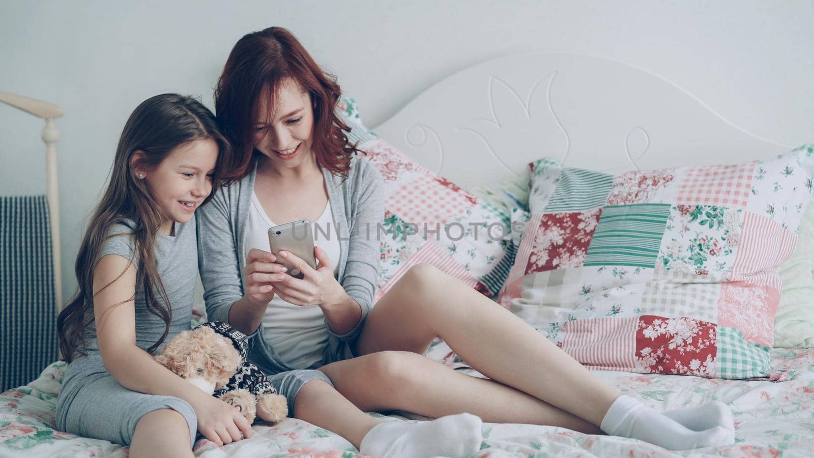 Smiling young mother with little cute daughter watching social media photos on smartphone using touchscreen and have fun while sitting in cozy bedroom at home by silverkblack