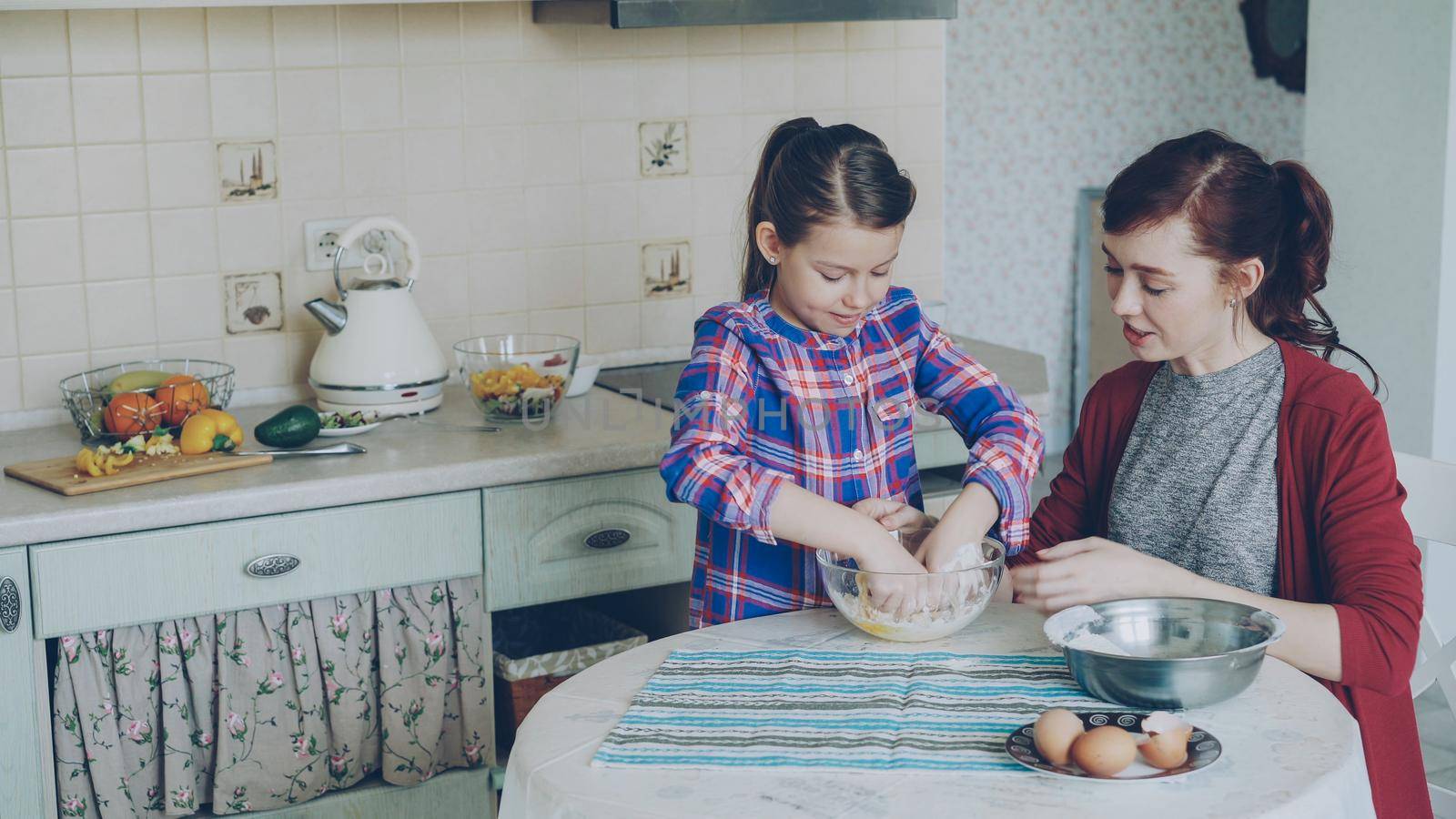 Little cheerful and funny girl helping her mother in the kitchen cropping egg into bowl and mixing dough for cookies. Family, food, home and people concept