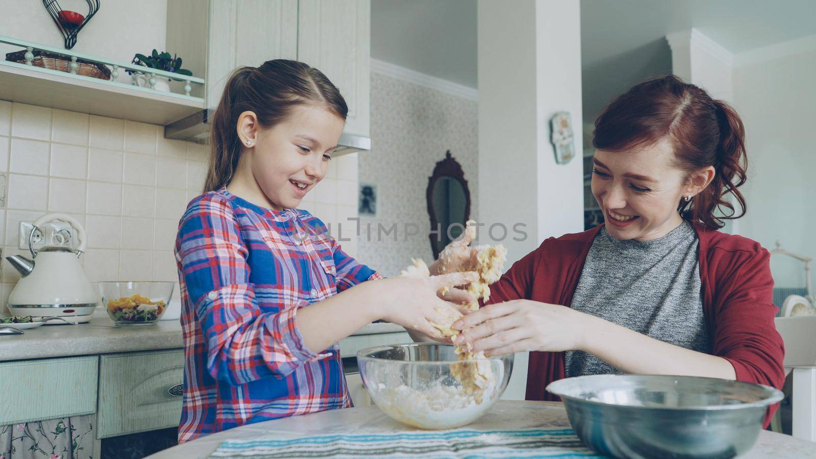 Happy young mother and beautiful daughter cooking together and having fun. Mom taking off sticky dough from girl hands and laughing together. Family, food, home and people concept