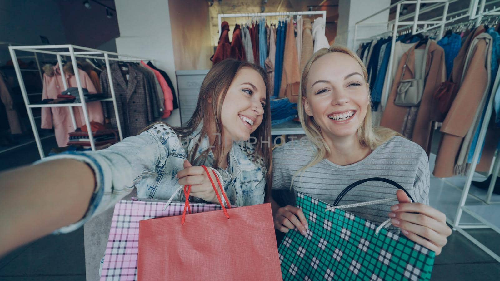 Point of view shot of two beautiful young women making selfie with paper bags in women's clothing department. Girls are posing, laughing and chatting happily by silverkblack