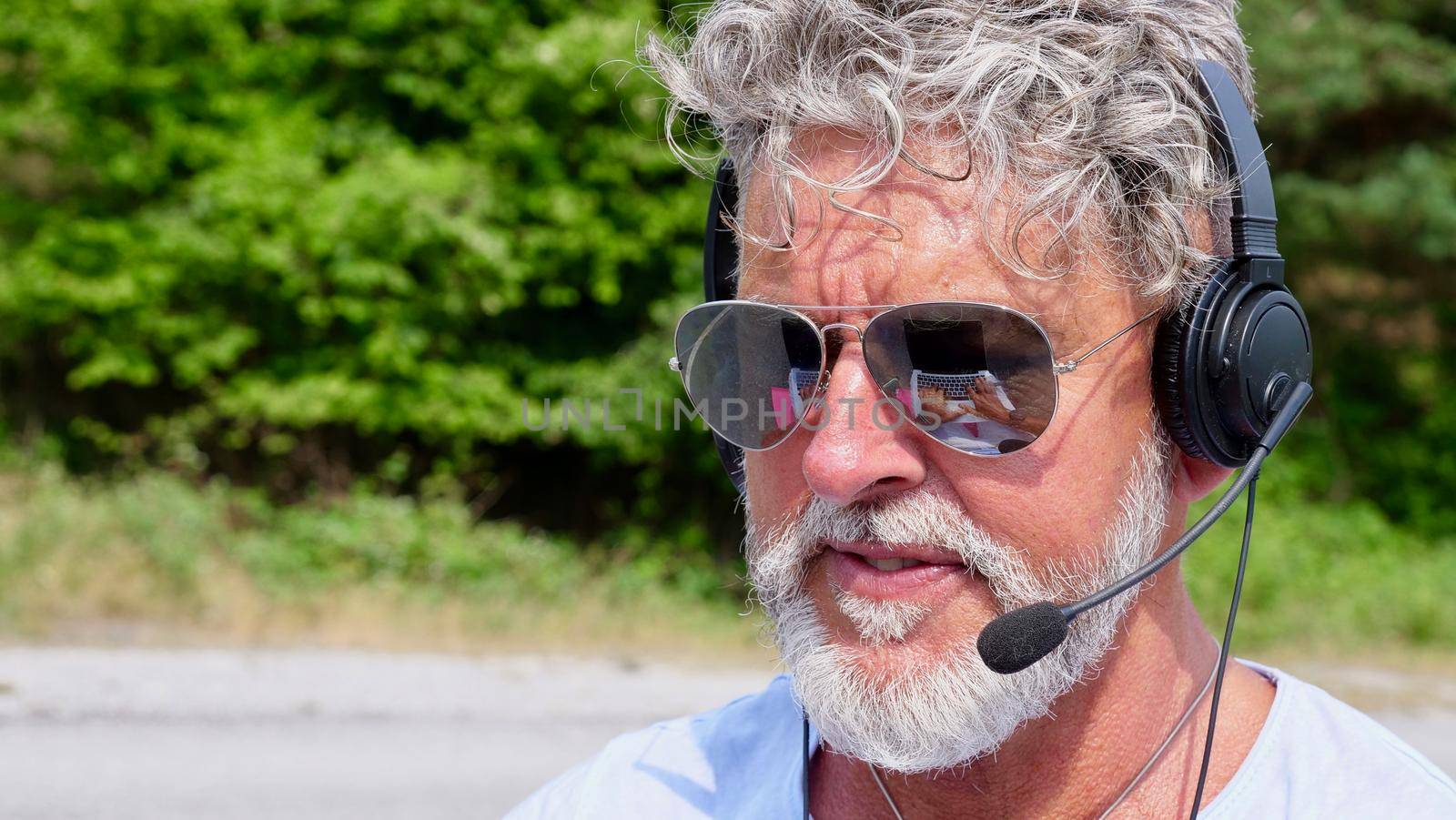 Gray-haired elderly man freelancer with a beard in sunglasses, working in the headphones on nature. A crazy and extraordinary old senior in the summer