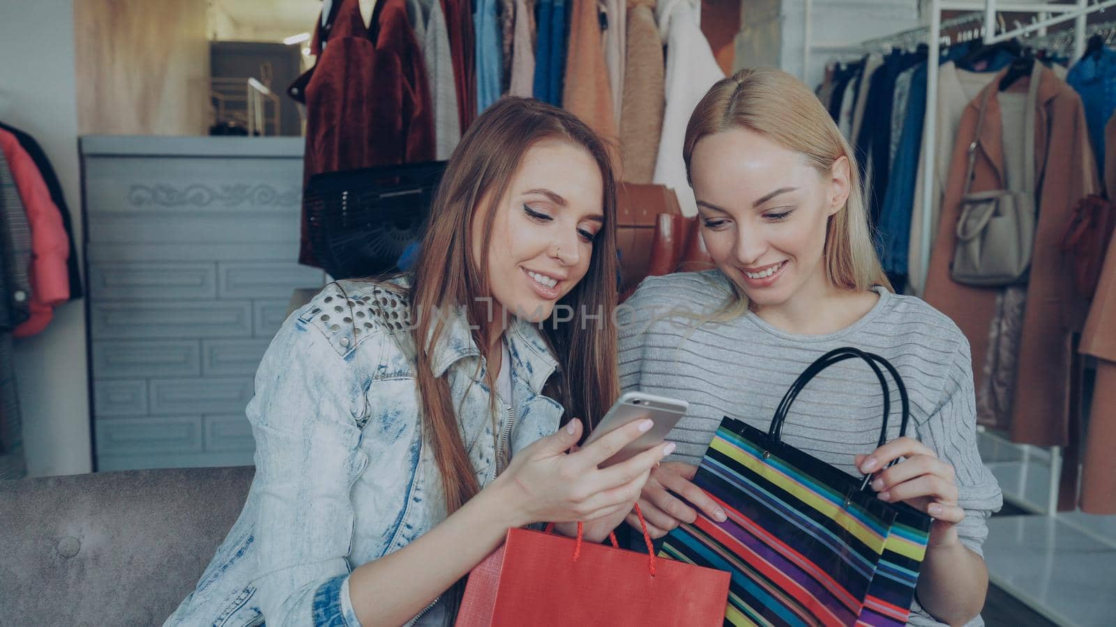 Attractive young ladies sitting on chairs in a clothing store with coffee and shopping bags, checking smartphone and talking. Shelves and hangers with colourful clothes in background