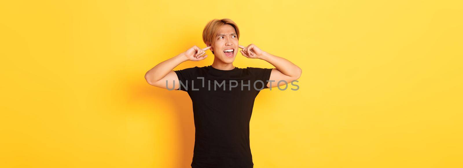 Portrait of annoyed and bothered asian guy grimacing, shut ears disturbed by loud noise, standing yellow background.