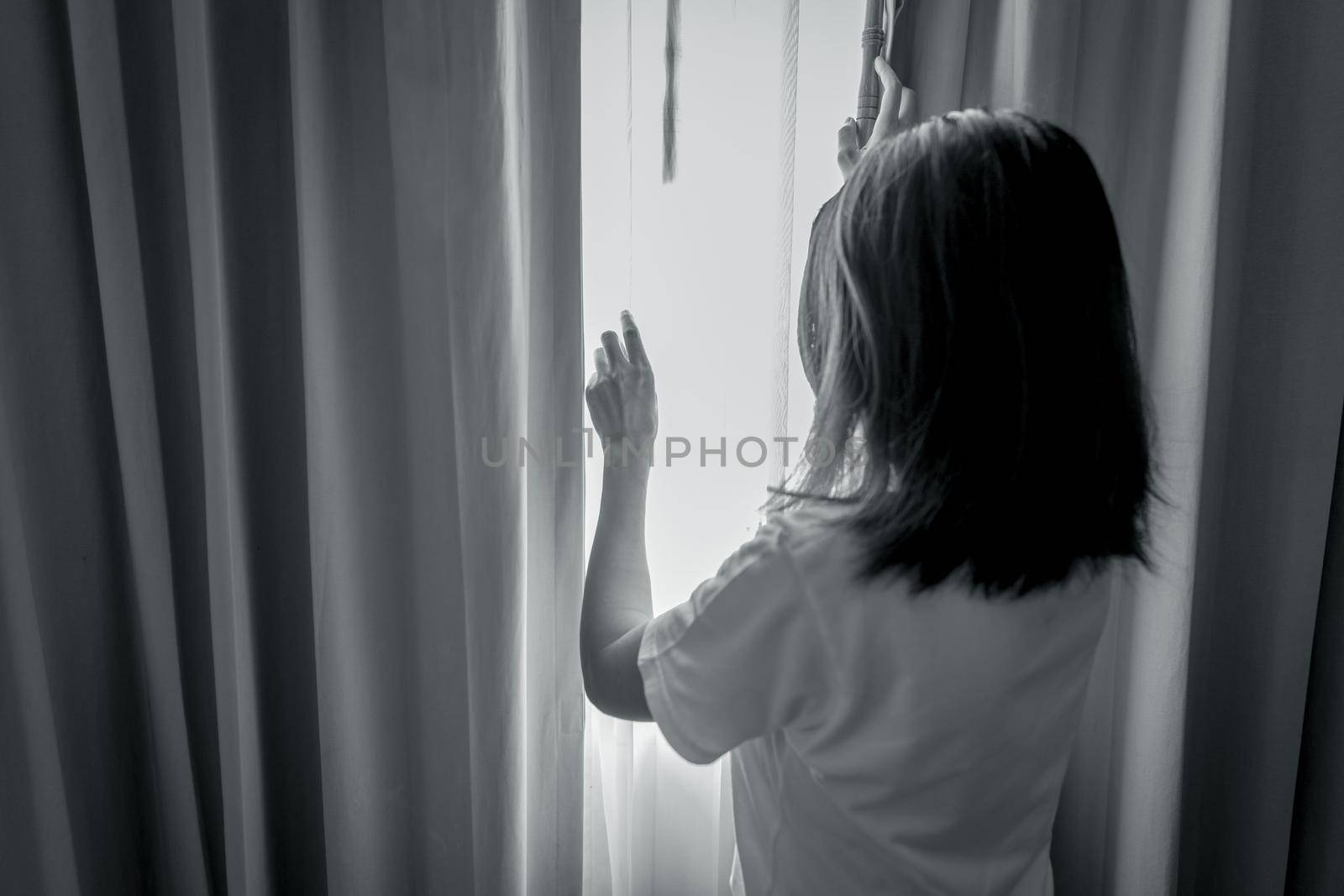Rear view of woman holding curtain and looking out of the bedroom. Depressed and stressed woman. World mental health day concept. Mental health awarenes. Thoughtful woman in dark room. Unhappy woman.  by Fahroni