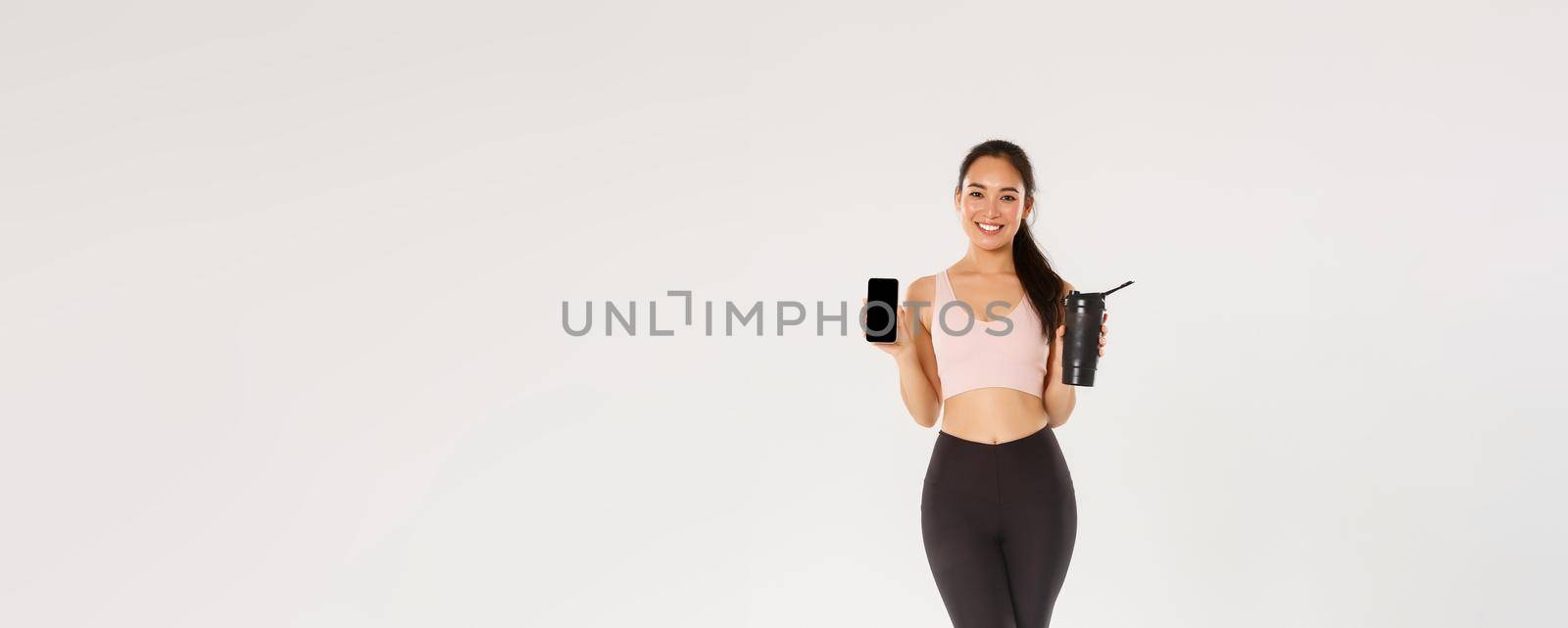 Full length of smiling good-looking asian fitness girl, sportswoman in activewear showing smartphone screen and water bottle, control drinking liquid with mobile application during training session by Benzoix