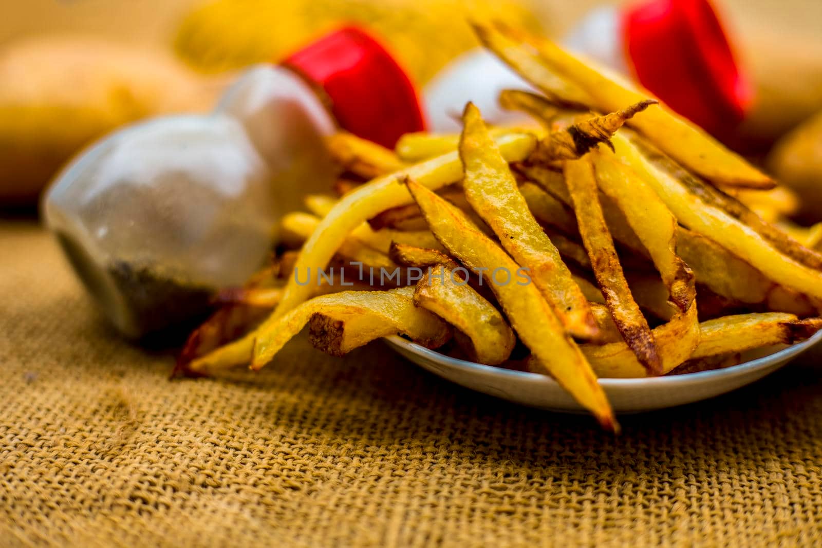Fresh fired French fries of along with sprinkler of black pepper and salt on jute bag's surface and some raw potato also.