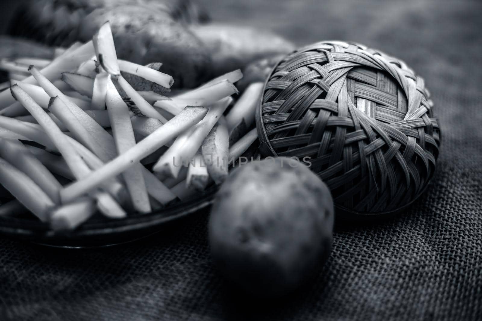 Raw cut french fries in a transparent glass plate along with raw potato with it on jute bag's surface.