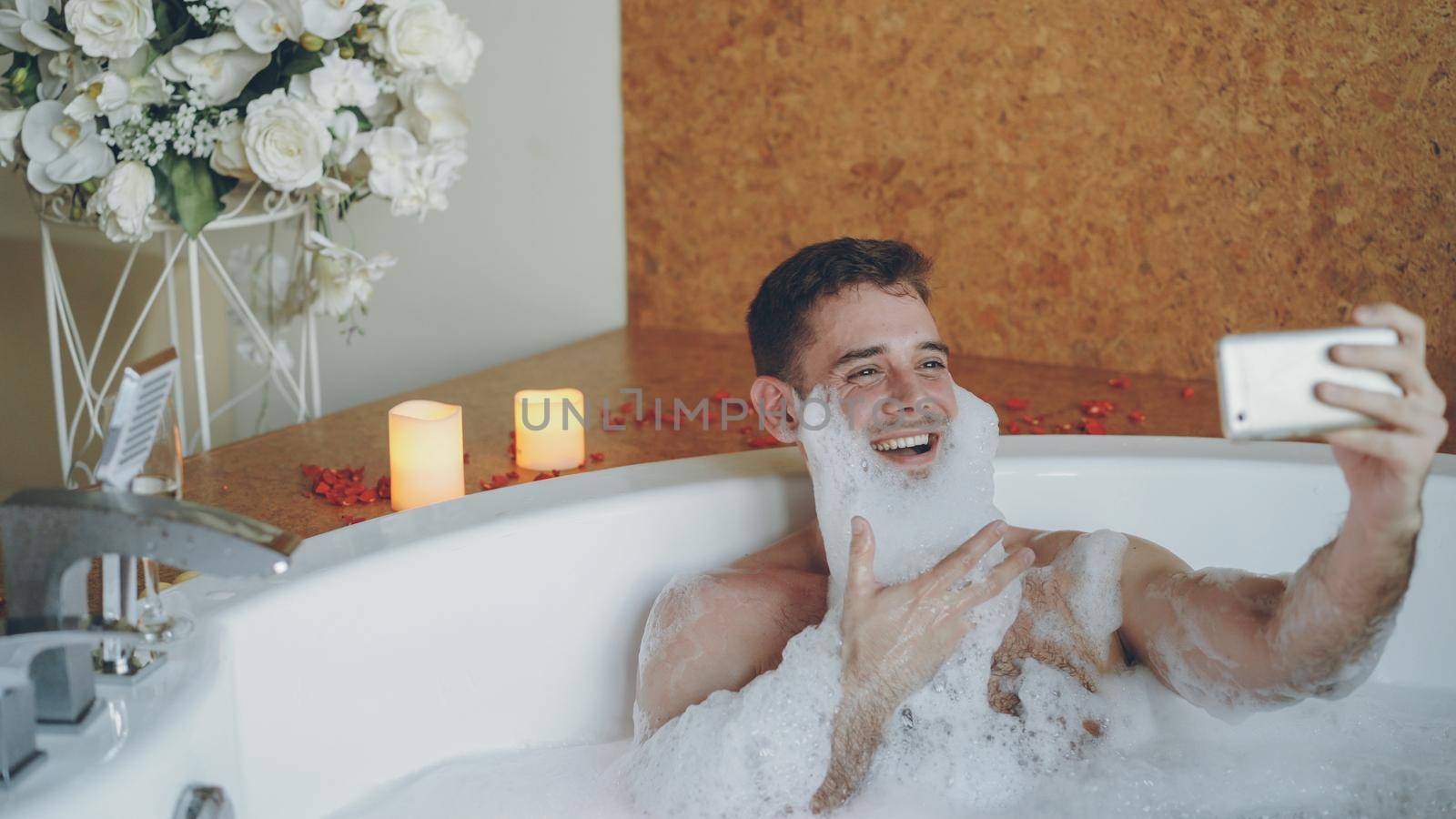 Cheerful young man with foam on beard is taking selfie using smartphone in hot tub in modern spa salon. He is laughing, gesturing, posing and looking at camera.